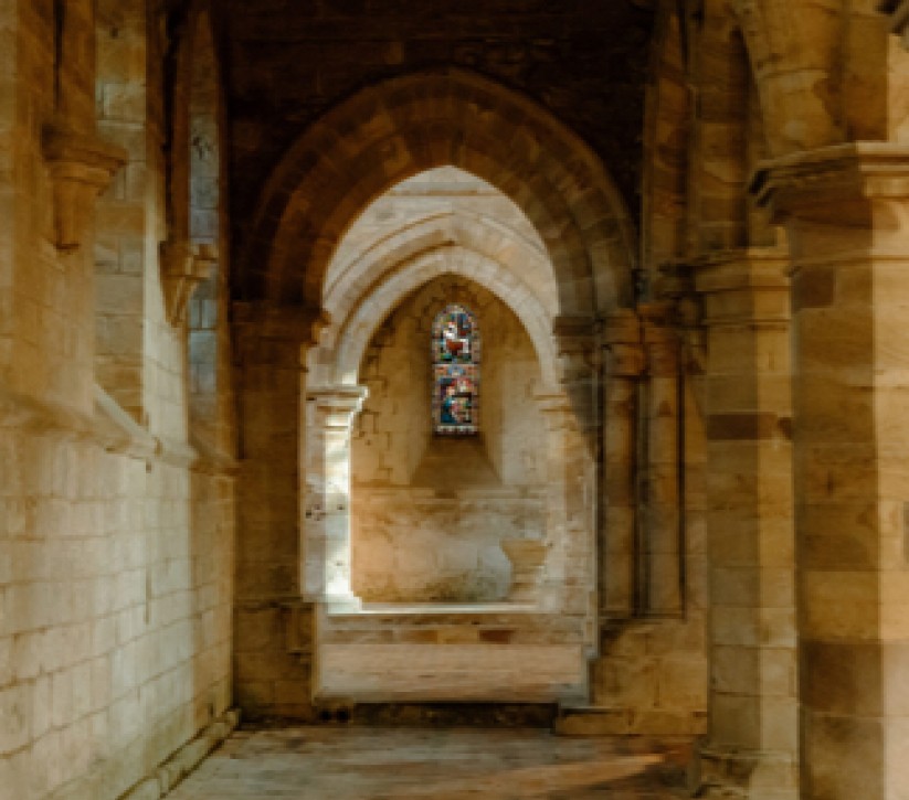 A small stained glass window at the edge of a grand church halllway